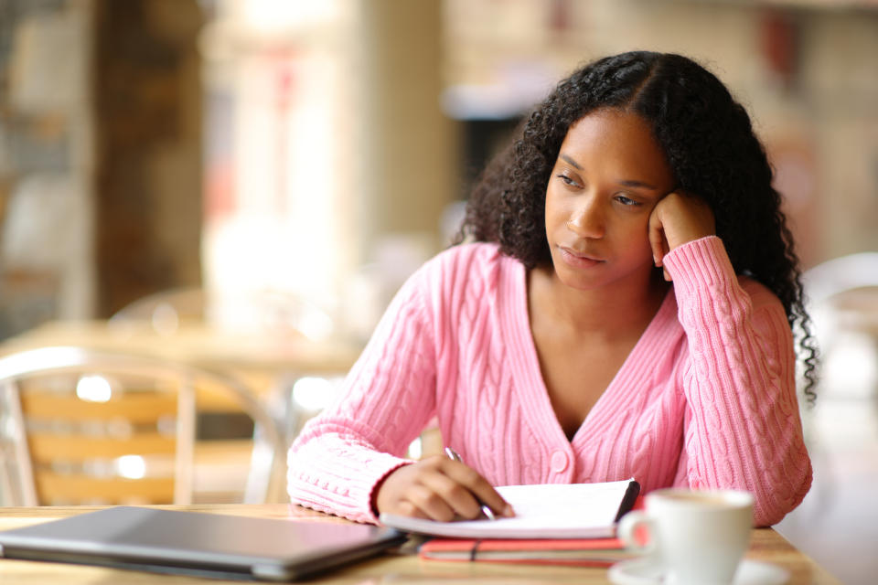 A person with long, curly hair wearing a cozy sweater is sitting at a table, looking thoughtful while writing in a notebook, with a laptop and coffee cup nearby