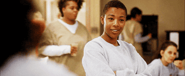 Samira Wiley in a casual setting, wearing a sweatshirt, smiles while crossing her arms. In the background, people in casual attire are visible