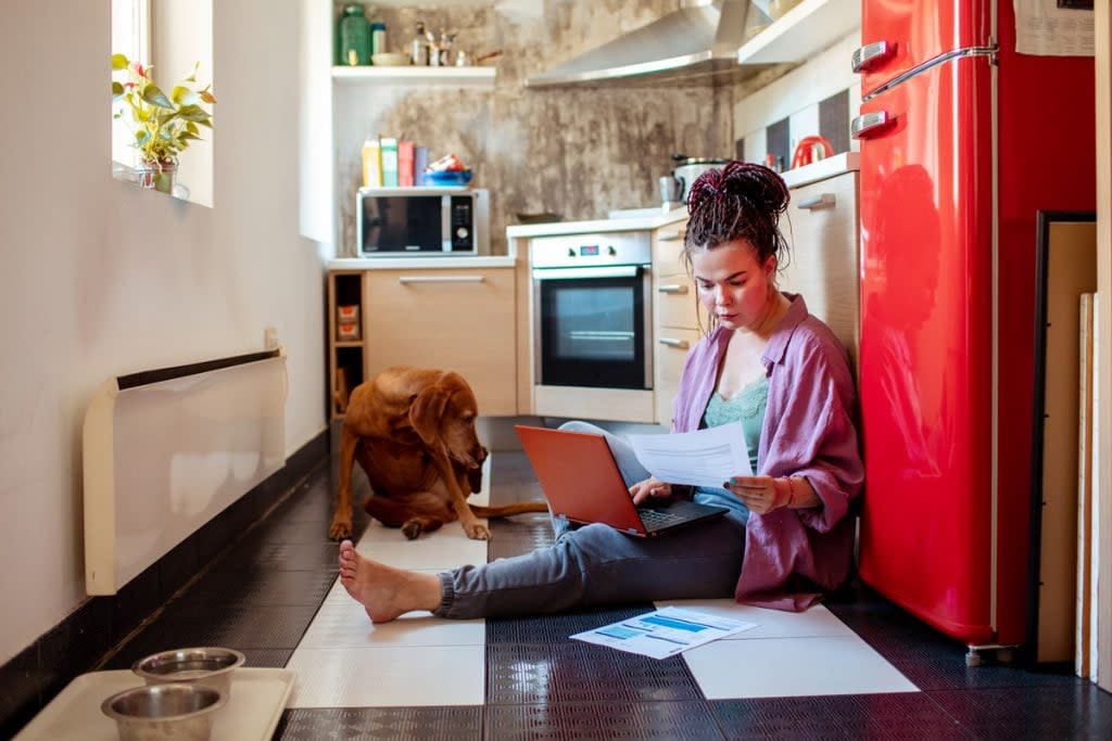 A woman uses her laptop while sitting on the kitchen floor to budget her monthly expenses. 