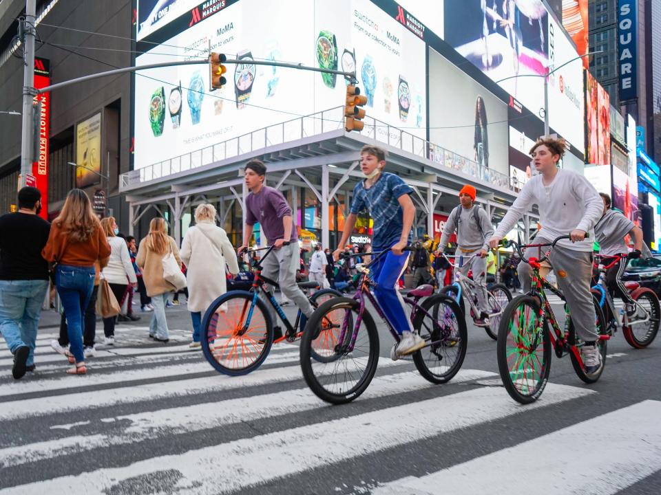 Three people ride bikes on a crosswalk in Times Square with bright screens displaying ads in the background.