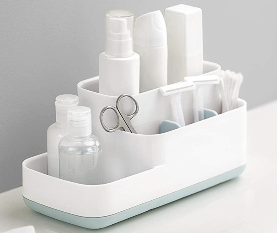 A white and pale blue Joseph Joseph EasyStore Bathroom Caddy pictured on a white bathroom surface against a grey wall, filled with razors, scissors, bottles etc.