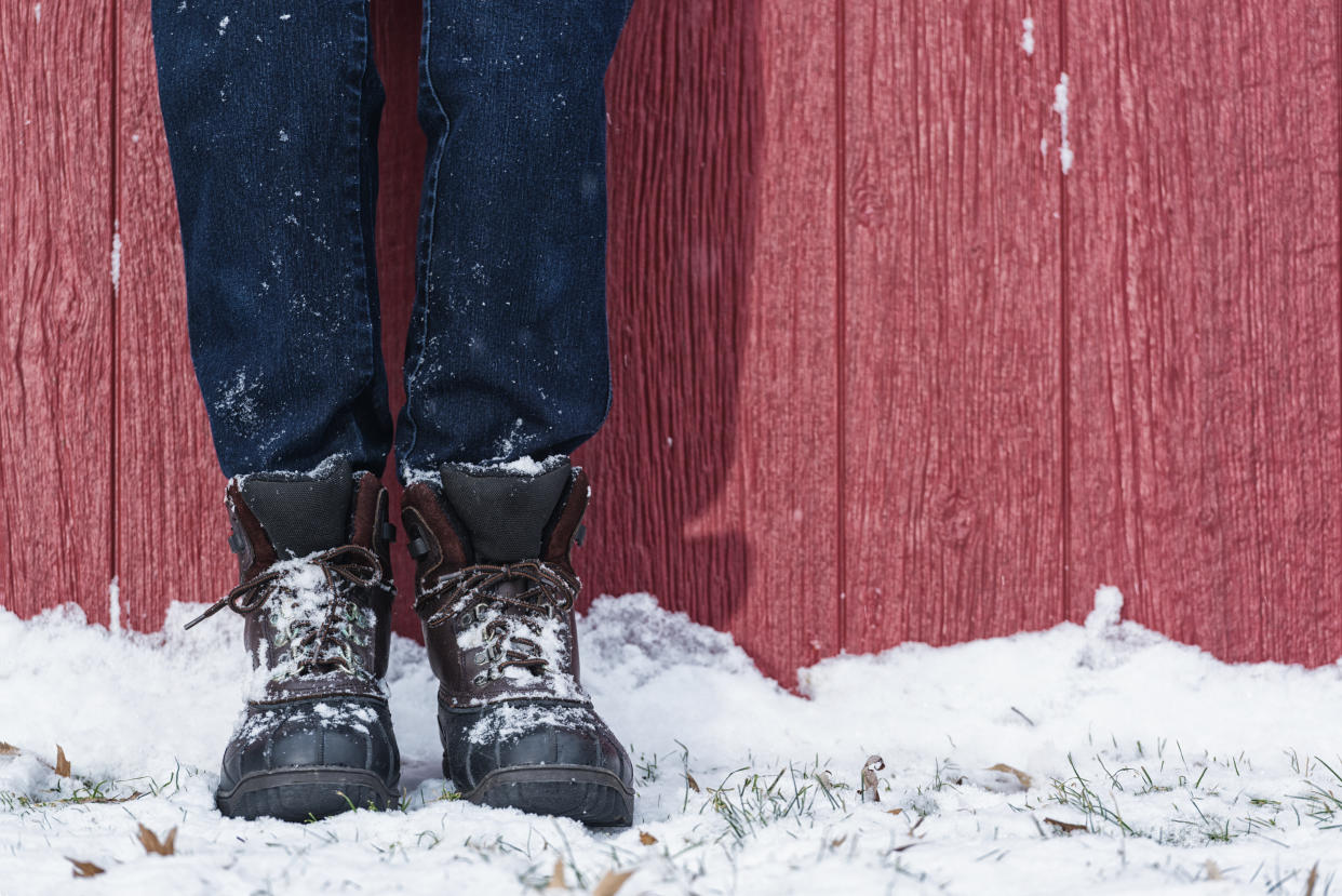 A close-up of a woman's legs wearing duck boots and blue jeans leaning against a red barn in the snow.