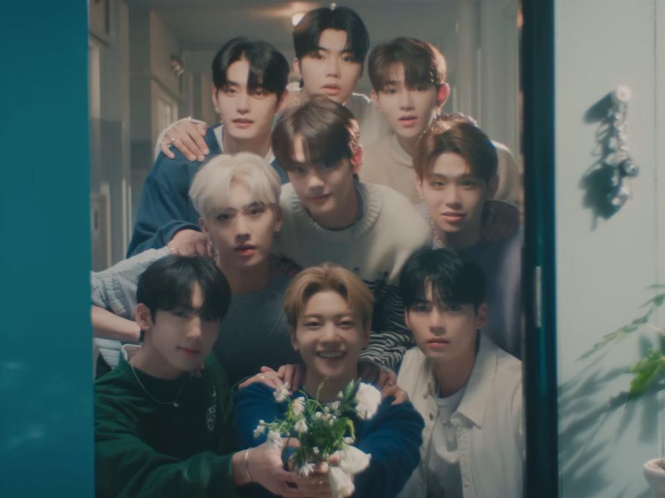 a group of nine young men standing in front of an open doorway. in the center, one is holidng out a bouquet of flowers and smiling widely