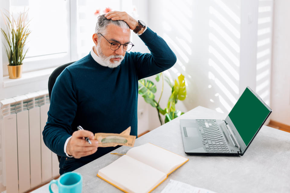 A mature Caucasian man is shown sitting at his desk at home, looking troubled and stressed as he goes through his paperwork, likely related to his bills