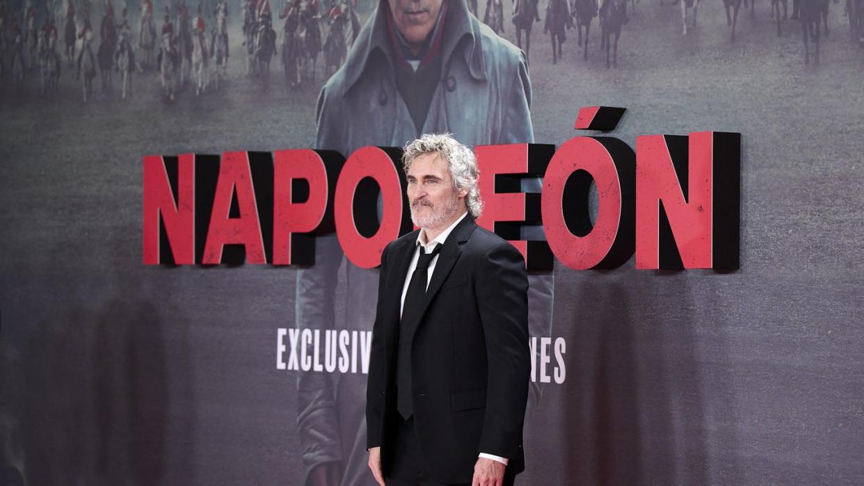 joaquin phoenix stands in a black suit in front of a large poster for the napoleon movie