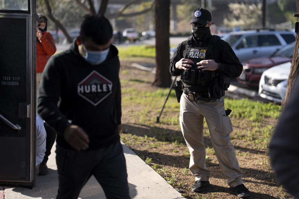 A Homeland Security officer looks on as migrants arrive at a bus stop after leaving a processing facility, Friday, Feb. 23, 2024, in San Diego. Hundreds of migrants were dropped off Friday at a sidewalk bus stop amid office parks in San Diego with notices to appear in immigration court after local government funding for a reception center ran out of money sooner than expected. (AP Photo/Gregory Bull)