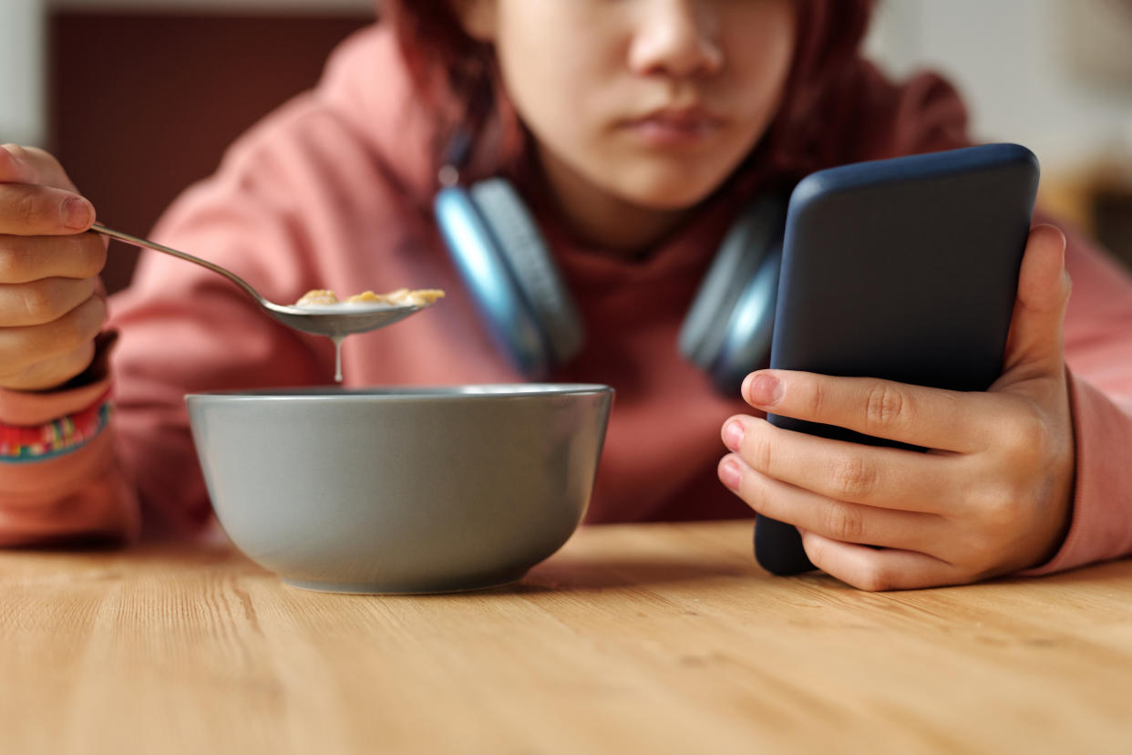 Hand of teenage girl with mobile phone having cornflakes with milk from bowl while sitting by table in front of camera and reading messages