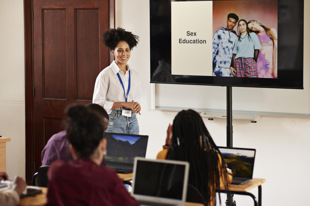 Smiling teacher standing by screen explaining sex education subject to students in classroom at high school