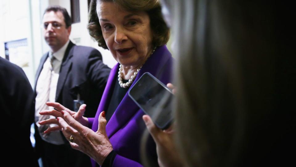 dianne feinstein, wearing a purple outfit, speaking to a reporter who uses her smartphone as a tape recorder