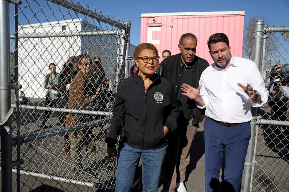 A woman and two men walk through a gate in a chain link fence with people in the background.