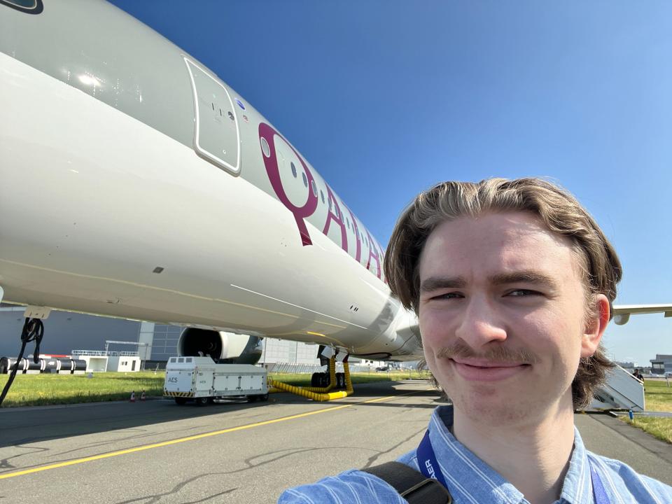 A young man in a blue shirt takes a selfie in front of a Qatar Airways Airbus A350