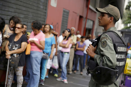 A Venezuelan soldier stands guard next to people forming a line to try to buy cornmeal flour and margarine at a pharmacy in Caracas March 15, 2016. REUTERS/Carlos Garcia Rawlins/File Photo