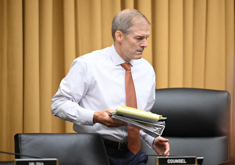 Jim Jordan stands in front of a yellow curtain next to a black chair holding a yellow notepad and white three-ring binder, among other papers.