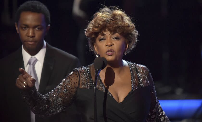 A woman with short hair wearing a black gown speaks and points onstage behind a microphone