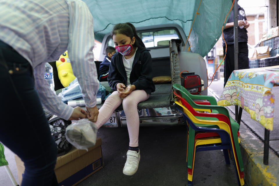 Neighborhood volunteer Rebeca Rodriguez ties plastic bags over the shoes of Paulina Mariano Ortiz who arrives for a lesson in the bed of a pick-up truck repurposed as an educational space on the southern edge of Mexico City, Friday, Sept. 4, 2020. Concerned about the educational difficulties facing school-age children during the coronavirus pandemic, the couple who runs "Tortillerias La Abuela," or Grandma's Tortilla Shop, adapted several spaces outside their shop to provide instruction and digital access to local children who don't have internet or TV service at home, a project that has attracted donations and a waiting list of students. (AP Photo/Rebecca Blackwell)
