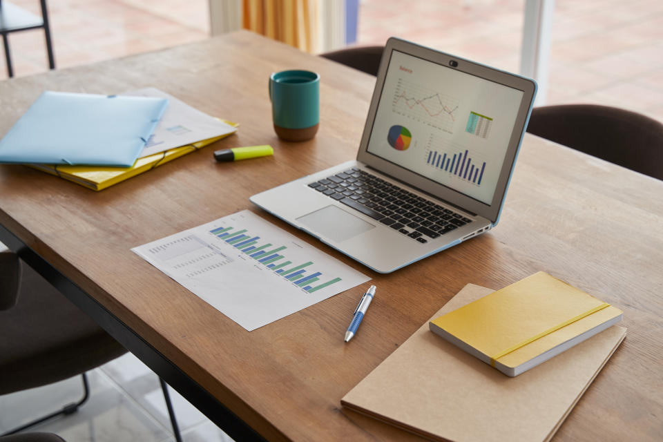 A laptop sitting on a desk with graphs open and journals and folders strewn around it