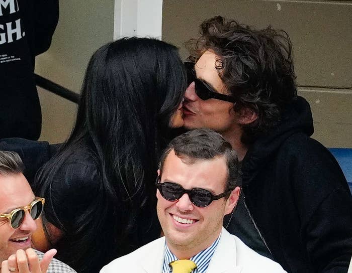 Kylie Jenner kissing Timothée Chalamet at an event, both wearing casual outfits. They are seated behind another man in sunglasses