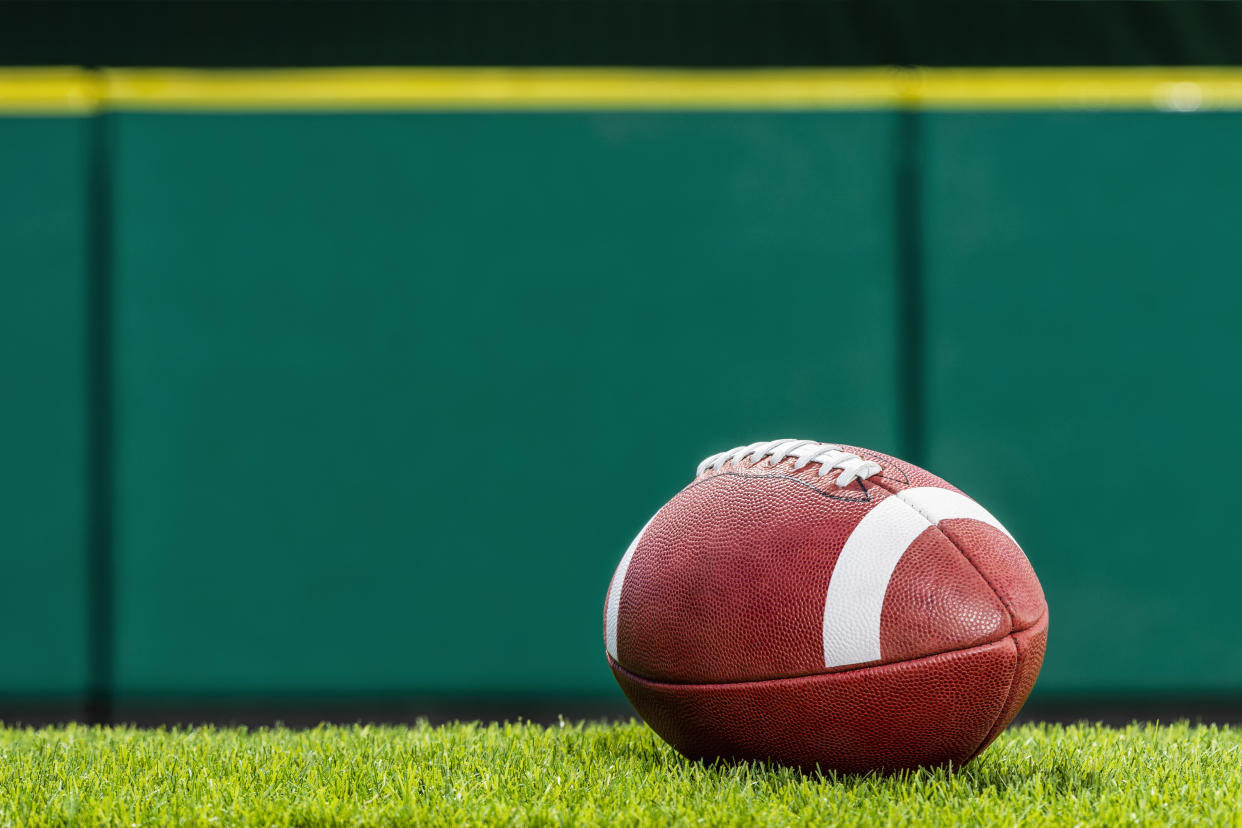 A low angle view, of a textured College or High School American Football made of leather with white stripe sitting on artificial turf of a stadium with green padded wall in the background. This type of football with the white stripes is use by colleges and high schools in the US.