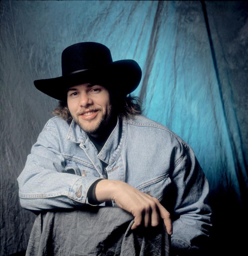 toby keith smiles at the camera while sitting in front of a background lit with a blue light, he wears a light colored jean jacket and a black cowboy hat