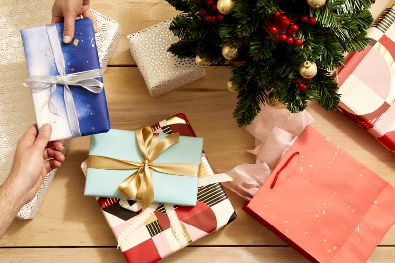 Overhead shot of someone handing over a gift wrapped in silver and blue wrapping paper, and tied in a silver bow, over a light wood surface.  On the light wood surface theres an assortment of other wrapped gifts and gift bags.