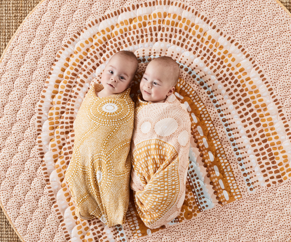 Two swaddled babies lay on the ground next to each other ontop of an indigenous rainbow play mat.