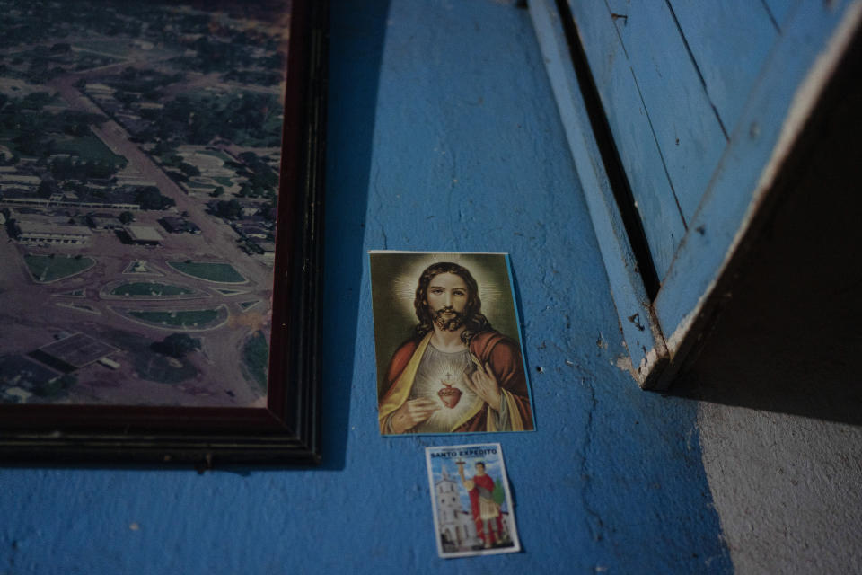 In this Nov. 29, 2019 photo, an image of sacred heart of Jesus hangs on a wall in Dede Diniz's home in the town of Ruropolis, Para state, Brazil, next to an aerial photo of the town. The Trans-Amazon highway and route BR-163 meet in the city of Ruropolis, where the military government promised land to lure people to the planned agricultural village in the 1970s. (AP Photo/Leo Correa)