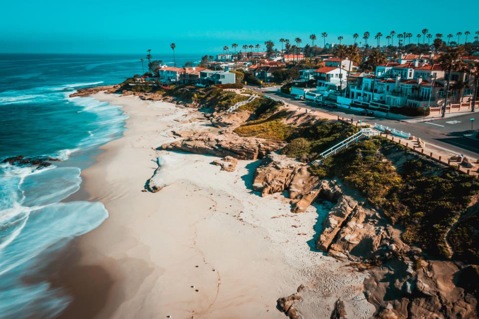 Aerial view of a coastal area with a sandy beach, waves, a road, and houses