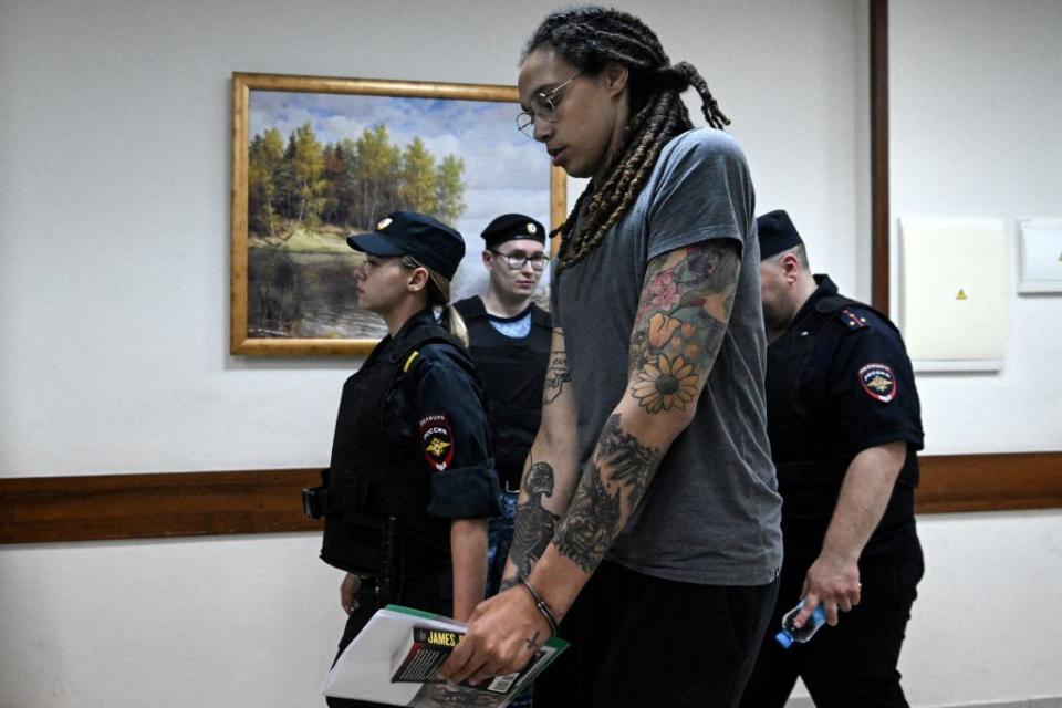 brittney griner, wearing glasses and a blue prison jumpsuit, is handcuffed and escorted down a hallway by uniformed officers