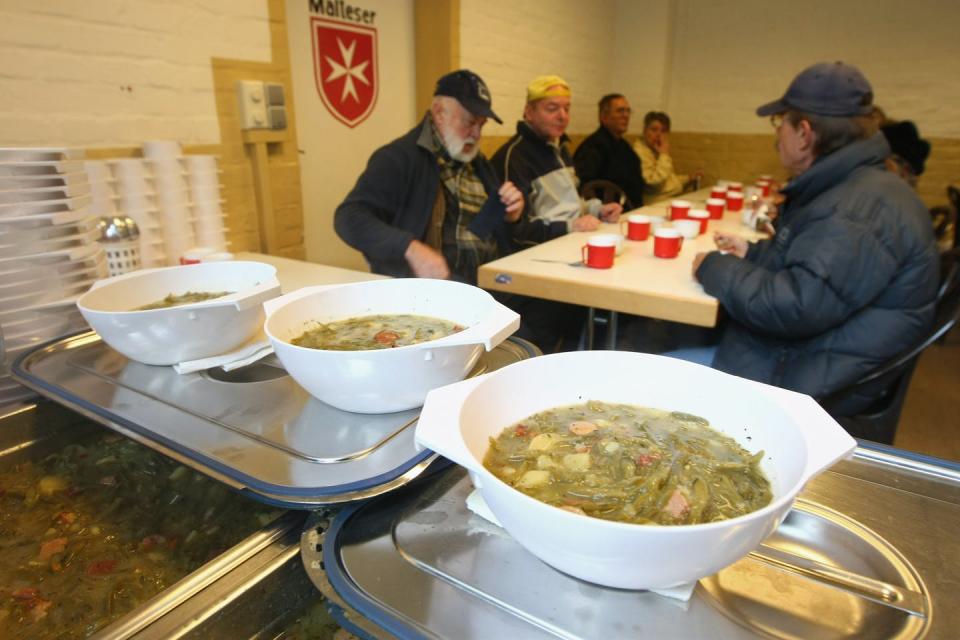 Older men sit around a communal table with red plastic cups. Bowls of hot soup can be seen in the foreground