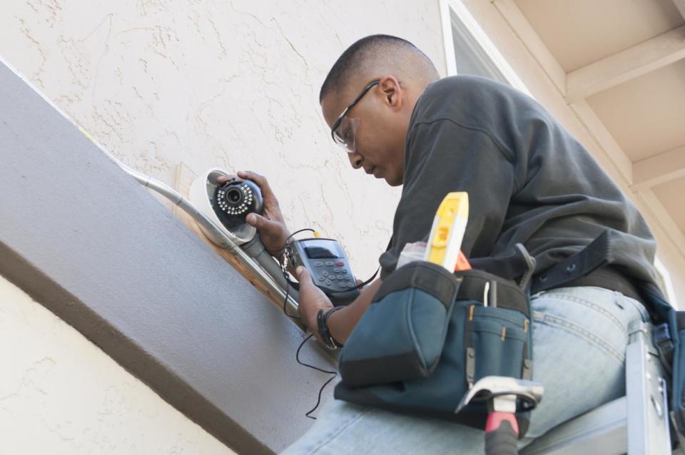 A worker stands on a ladder to install a security camera.