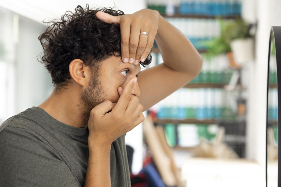 Side view medium shot a young male adult applying a contact lens looking in a vanity mirror wearing casual clothing.