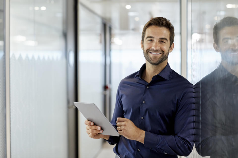 Portrait of smiling businessman holding digital tablet. Mid adult male professional is wearing formals. He is leaning on glass at corporate office.