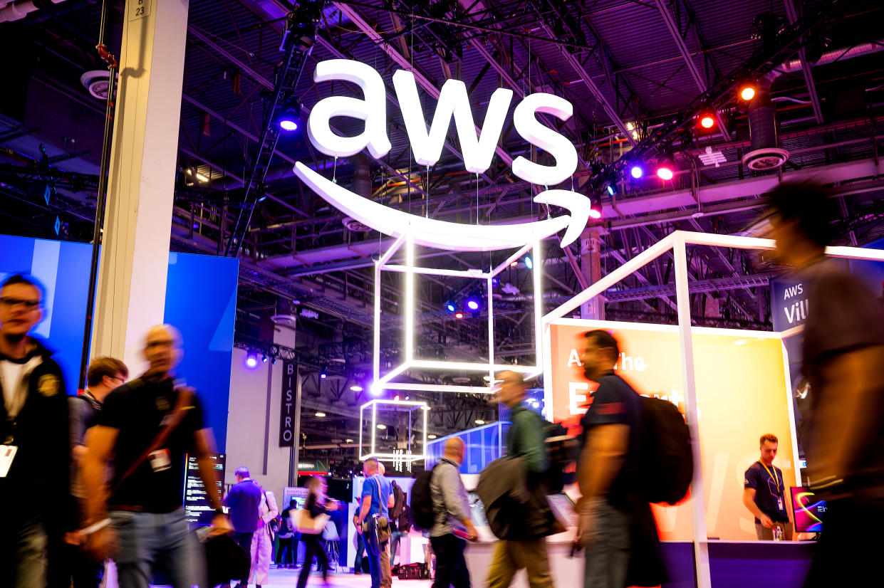 People in motion walk around an expo hall under a lit up AWS sign hanging from the ceiling.