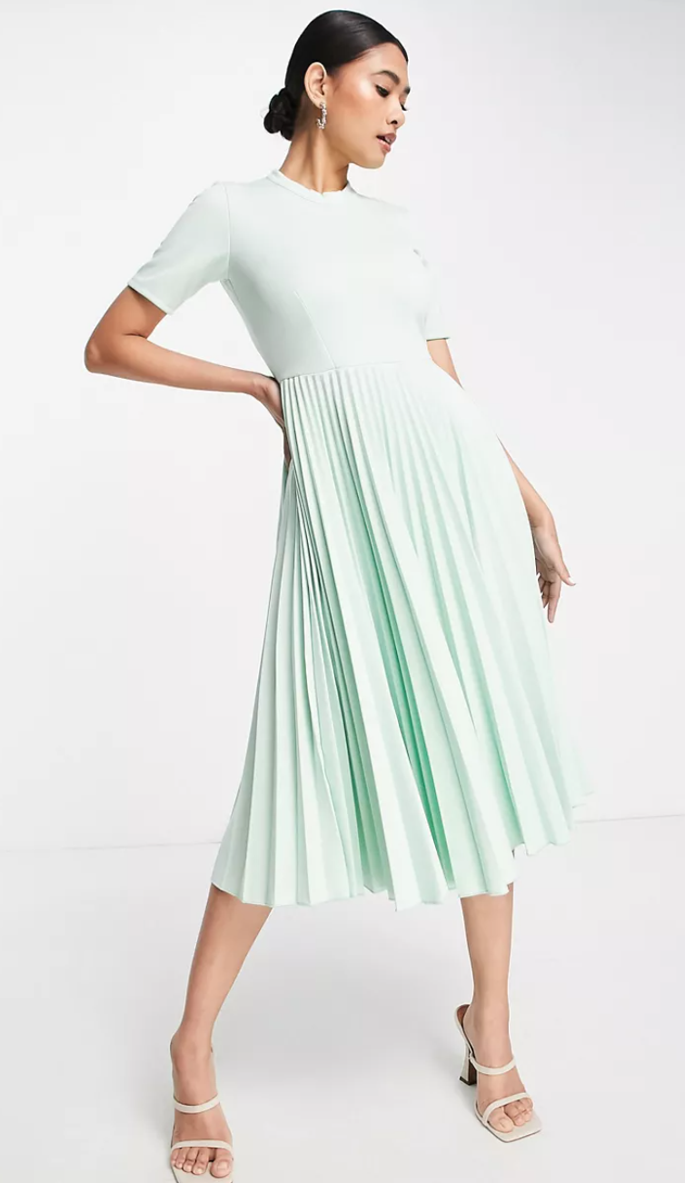 A model shows off a pale green dress with short sleeves and midi pleated skirt. She stands with a hand on one hip against a pale background.