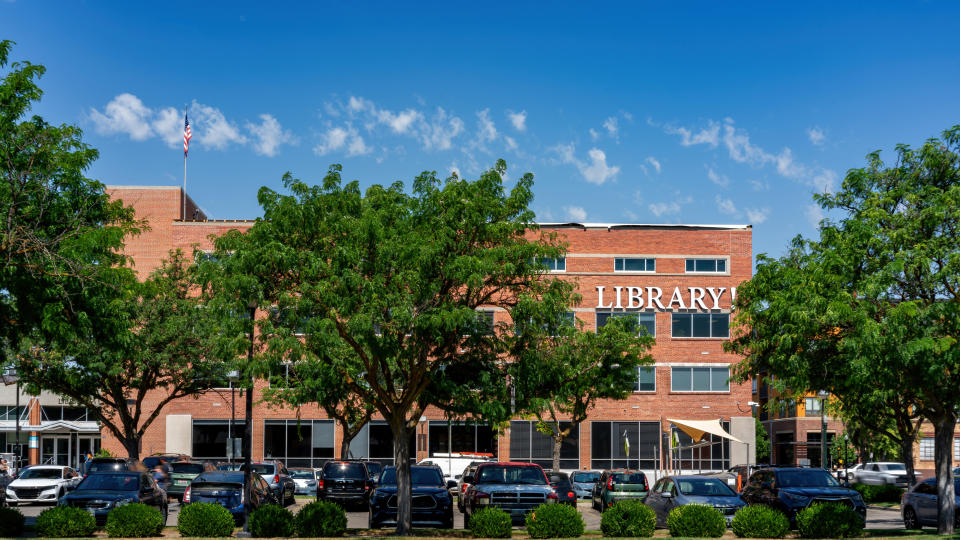 A public library with a brick exterior, trees in the foreground, and parked cars in front of the building