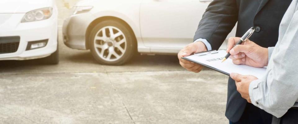 two men up close, holding a clipboard and pen, two cars in the background