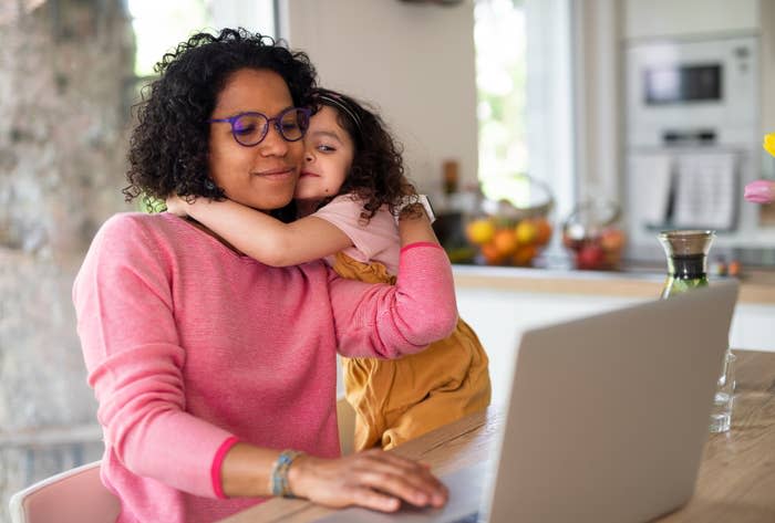 Woman at laptop with child hugging her from behind, both smiling, in a kitchen setting