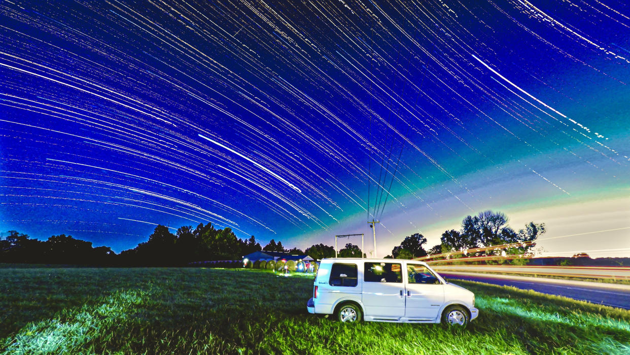  A white van is parked in a grassy field next to a road, lined with the streaking lights of a passing car. the sky is filled with the trails of stars. 