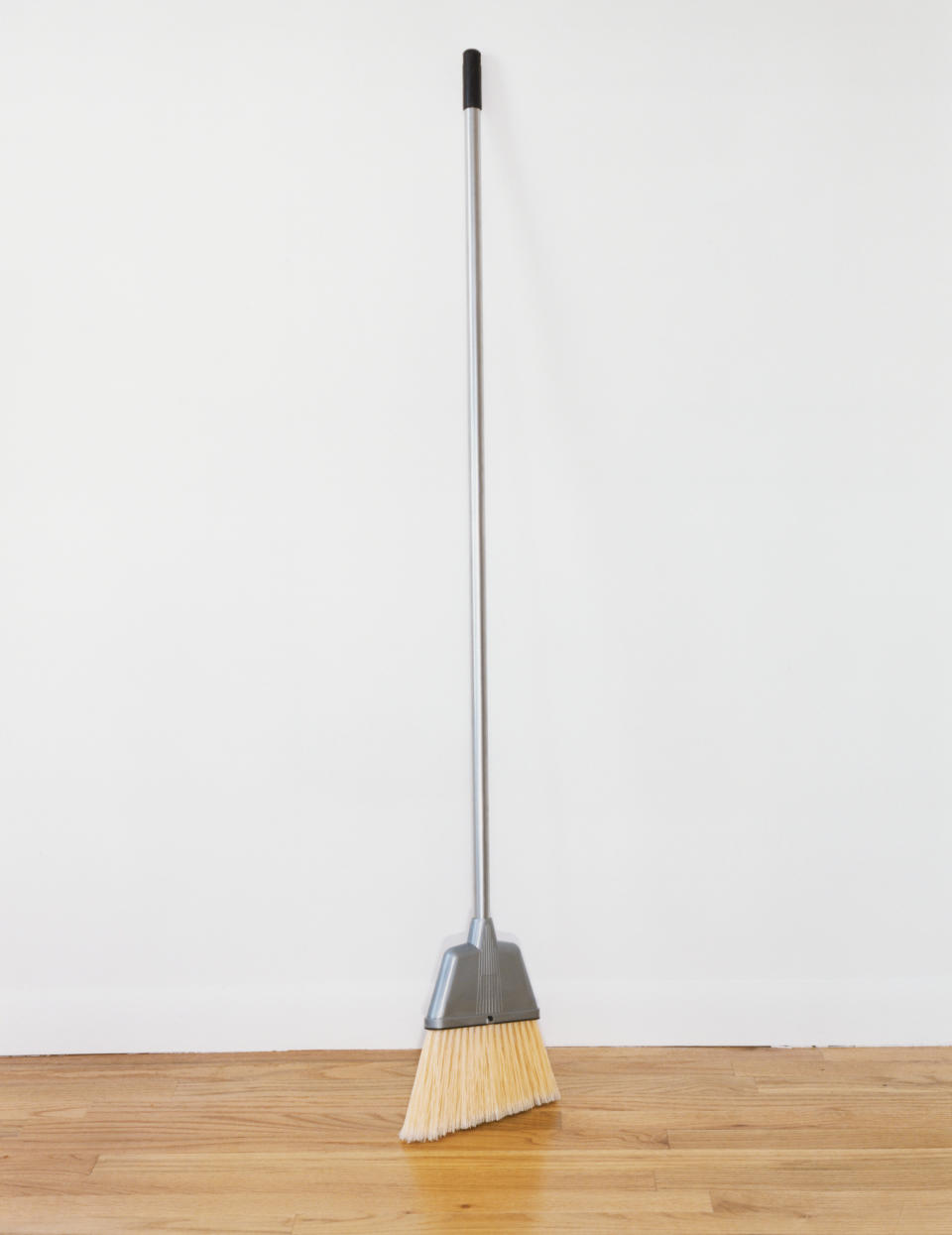 Broom with a silver handle and yellow bristles stands upright against a clean, white wall on a wooden floor