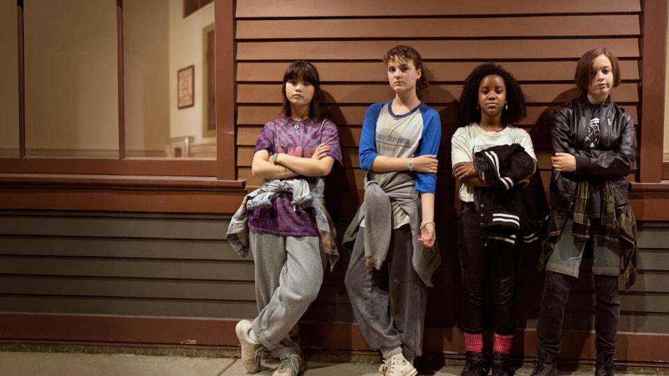 A promotional image from Paper Girls, showing on Amazon Prime Video from July 29, shows four female actors standing against a wooden wall staring at the camera wearing casual clothing.