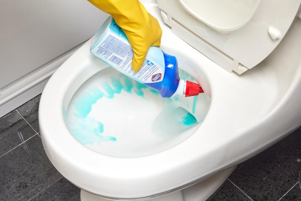 Woman wearing rubber gloves spraying toilet cleaner inside the rim of the toilet.