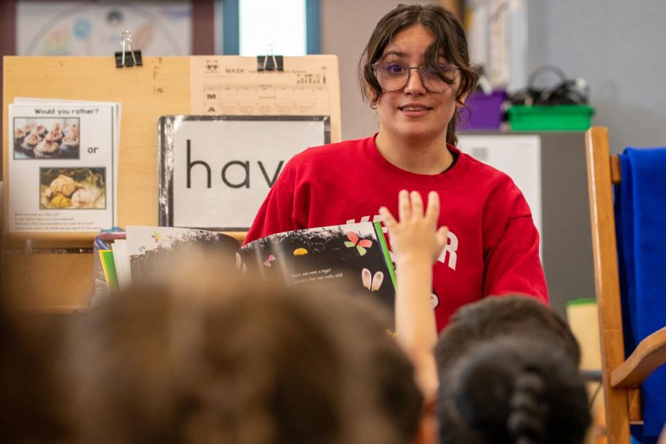 A seated woman reads to children in a classroom as one raises a hand.