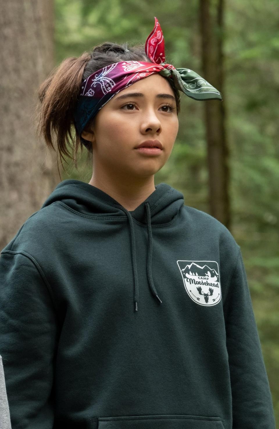 Dawn wearing a hoodie with a forest emblem, standing in a wooded area