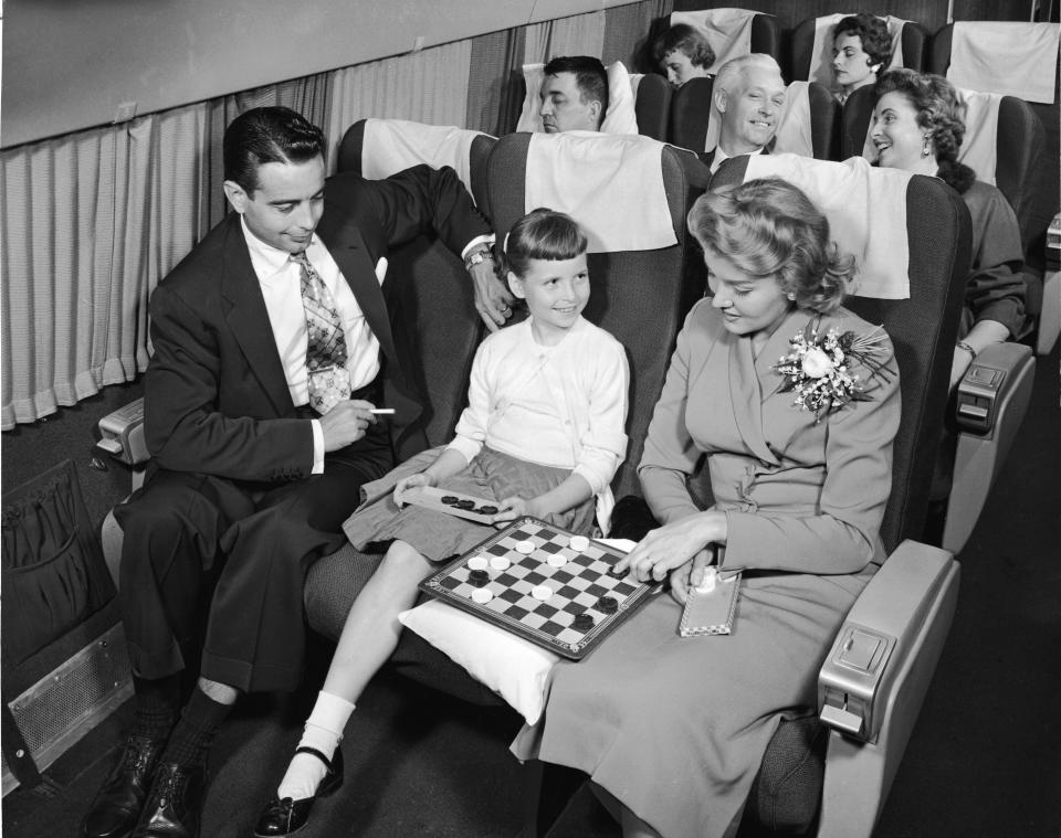 In this interior view of a commercial passenger plane in the 1950s, a woman contemplates a move in a checkers game she is losing to a young girl, while a man with a cigarette watches them.