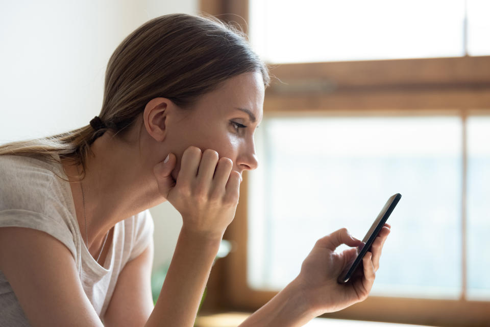 Close up side view face pensive sad young woman sitting near window holding smartphone absorbed reading unpleasant news, check social media messages, waiting sms from boyfriend date invitation concept