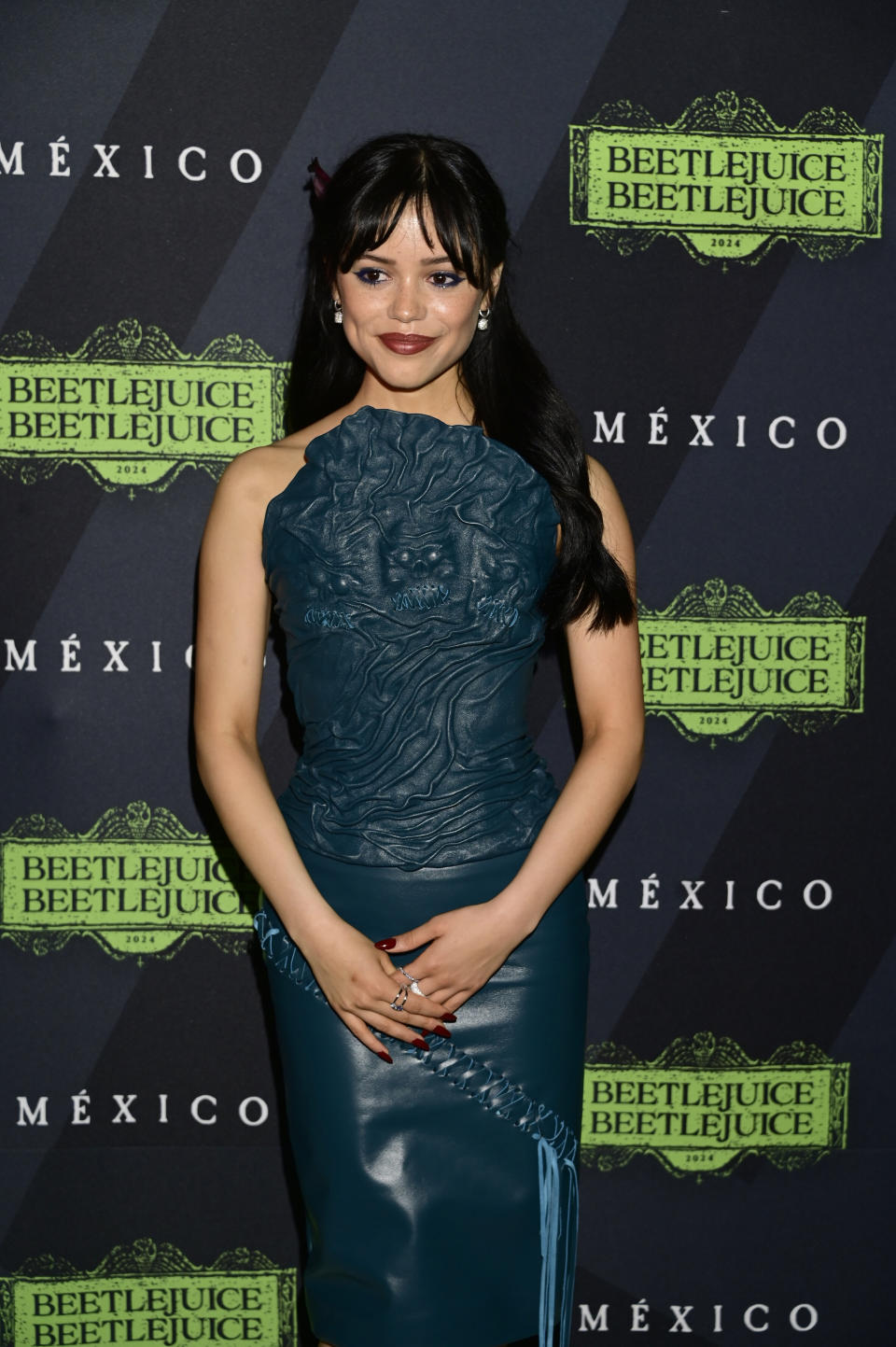 Jenna Ortega on the red carpet, wearing a stylishly sculpted blue dress, standing in front of a "Beetlejuice" movie promotional background with Mexico signage