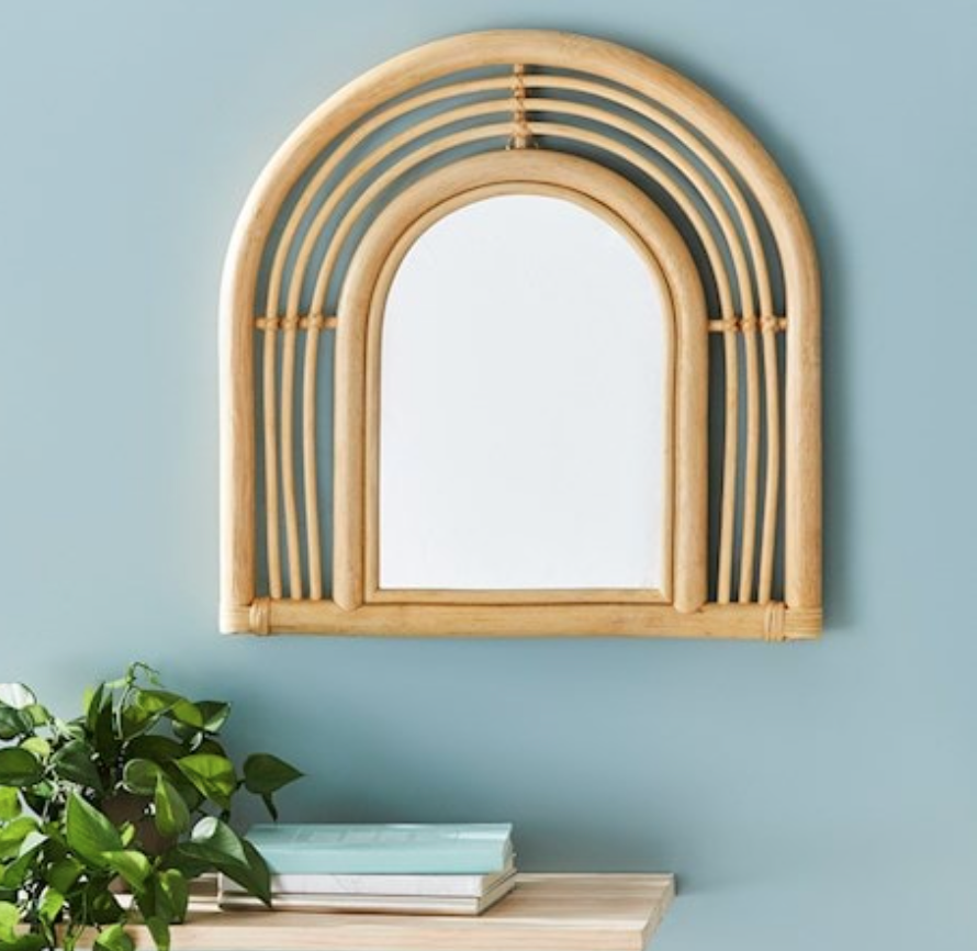 Kids' arch mirror in natural rattan hangs on a blue wall, above a sideboard holding books and a plant