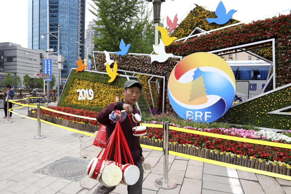 A man selling Chinese drums walk past a decoration promoting the upcoming Road and Belt Forum in Beijing on Friday, April 19, 2019. China is downplaying the political implications of its global development campaign known as the Belt and Road initiative, saying that it aims to boost multilateralism amid protectionist trends in the U.S. and elsewhere. (AP Photo/Ng Han Guan)