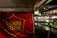 Tourists dine in restaurants at Whalers Village shopping mall in front of a "Lahaina Strong" flag from a housing protest on Kaanapali Beach, Wednesday, Dec. 6, 2023, in Lahaina, Hawaii. A group of survivors is camping on the resort beach to protest and raise awareness for better long-term housing options for those displaced. Residents and survivors still dealing with the aftermath of the August wildfires in Lahaina have mixed feelings as tourists begin to return to the west side of Maui, staying in hotels still housing some displaced residents. (AP Photo/Lindsey Wasson)