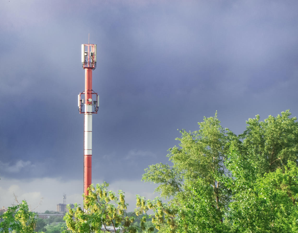 High tower of the GSM transmitter station in the evening against the background of blue sky and trees, in summer, after rain.